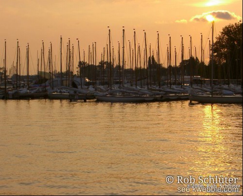 De zon gaat onder achter een rij masten van zeilboten.