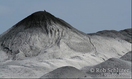 Klein silhouet van een kraai boven op een grote berg grijs zand.