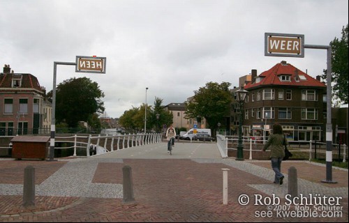 Vlakke fietsbrug met aan weerszijden borden op palen, een met 'heen', de ander met 'weer'.