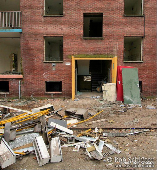 Gaten van ramen en deuren in de muur van een flat die gesloopt wordt tonen de lege ruimtes terwijl voor op straat het puin ligt.