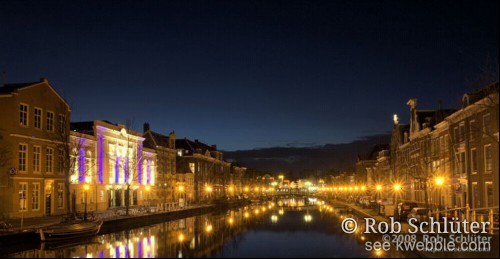 Een singel verlicht door het oranje licht van lantaarns, met op de linkeroever een wit gebouw dat blauwe strepen krijgt door gekleurde spots.