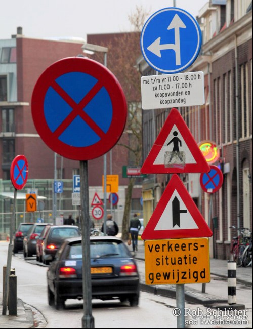 Straat met auto's en er langs een grote verzameling verkeersborden. Aan 1 paal hangen 5 borden die de rijrichting aangeven, laten weten wanneer die geldig is, waarschuwen voor overstekende voetgangers en een zijweg plus de mededeling dat de verkeerssituatie gewijzigd is.