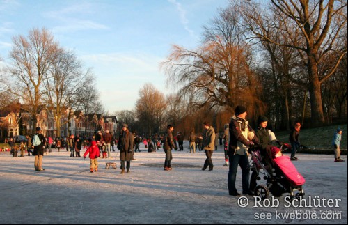 Mensen schaatsen, wandelen met een baby in een buggy en sleeën op een bevroren singel omzoomt door bomen.