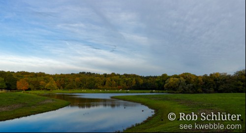 Een meertje slingert van links naar rechts door weilanden die lopen tot een bosrand in herstkleuren.