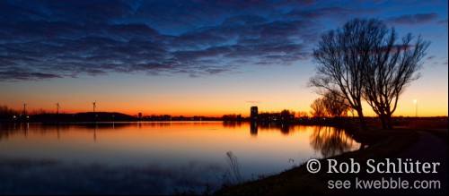 Avondopname met het vlakke water van het Spaarne met daarachter, van links naar rechts, de windmolens op Schoteroog, de flats van de wijk Land in Zicht en kale bomen langs het water. Hierboven een helderoranje gloed van de zon die reflecteert in het water en oplossende donkere wolkjes.