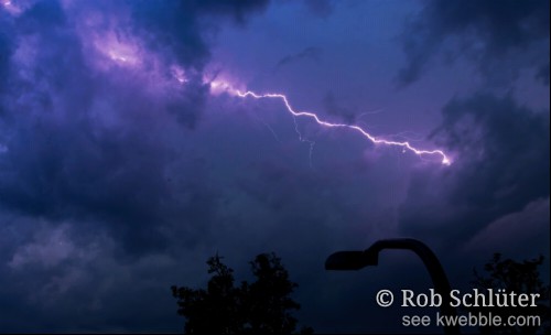 Een felwitte bliksemschicht in de lucht tussen donkerpaarse wolken met op de voorgrond een silhouet van de lichtkap van een lantaarnpaal.