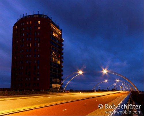 Onder blauwe avondwolken staat een bakstenen flat langs de Schoterbrug, een lange brug met apart fietspad. De weg wordt verlicht door lampen boven de weg aan metalen bogen.