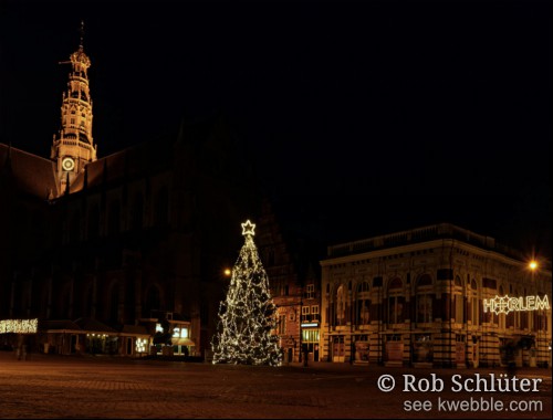 Het plein van de Grote Markt in Haarlem 's avonds in het donker, met een kerstboom met lampjes. Daarachter staan de Sint-Bavokerk met verlichte toren en andere oude gebouwen zoals de Vleeshal. Rechts hangt boven de straat de naam Haarlem in kerstverlichting.