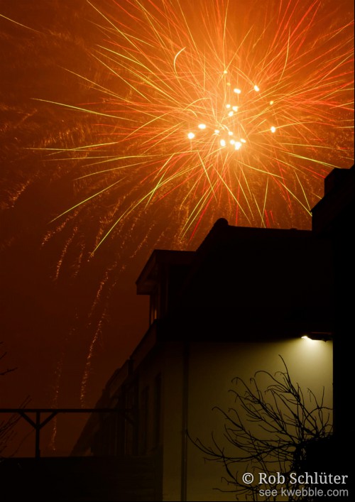 Een donkere dakkapel boven een muur in het licht van een straatlantaarn. Daarboven zetten rode en gele sporen van explodered vuurwerk de lucht in een oranje gloed.