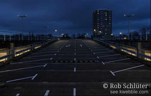 Donker en bewolkt, na zonsondergang maar nog geen nacht, op de lege bovenste verdieping van een parkeergarage. Je kijkt je uit over de brede toegangsweg met aan de zijkanten lege parkeervakken, die vanaf de lagere etage omhoog loopt naar het parkeerdek. In de achtergrond staat een torenflat met enkele verlichte ramen.