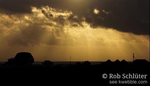 Gebouwen op de boulevard, waarvan rechts 3 strakke driehoeken, steken zwart af tegen de oranjegele lucht met zonnestralen die tussen de wolken door schijnen.