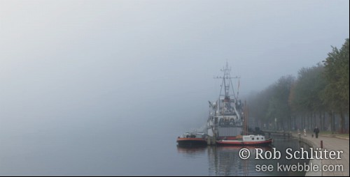 Weids uitzicht op mist die onzichtbaar in water overgaat. Een paar kleine boten en een fregat liggen dicht bij de kade, waar een donker geklede man met hond langs een rij bomen wandelt.